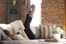 a woman lying on a couch reading a book with her feet in the air 