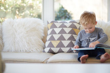 a toddler boy sitting on a couch reading a book 