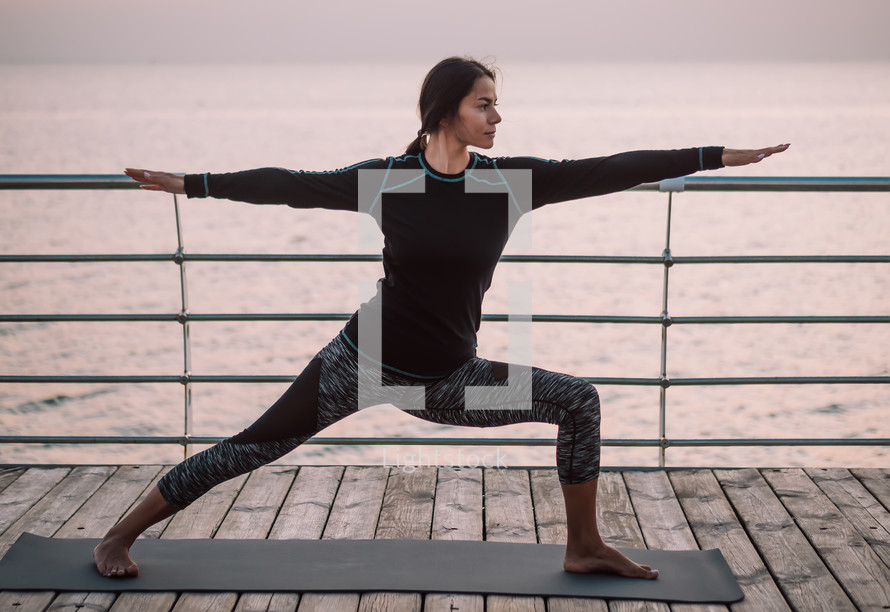 Young brunette hispanic woman doing yoga exercises on wooden sea embankment in the morning. Girl in black sports costume. Stretching, practice, healthy lifestyle concept .