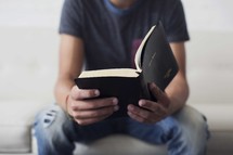 a teen boy reading a Bible on a couch.