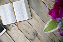cellphone, open Bible, and flowers in a vase on a wood table 