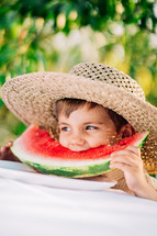 Cute little boy eating slice of juicy watermelon sitting on natural green garden background. Adorable kid in straw hat enjoying summer fresh melon fruit with smile. High quality photo