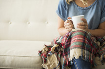 a woman sitting on a couch with a plaid blanket in her lap holding a mug 