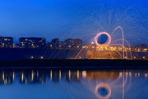 Hot Golden Sparks Flying from Man Spinning Burning Steel Wool near River with Water Reflection. Long Exposure Photography using Steel Wool Burning