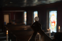 Hands holding up a communion wafer with stained glass windows in the background.