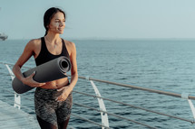Young sports woman walking to do morning stretching practice. Girl holding yoga mat. Sea or ocean background. Summer days. 