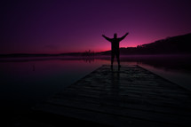 A man stands on a dock with his hands raised to the heavens.