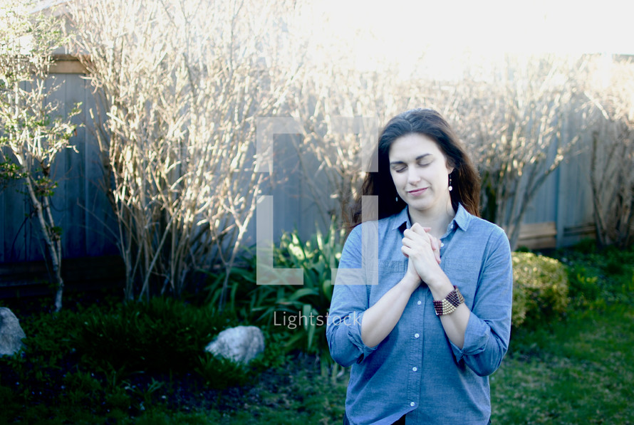 A woman standing outside in a garden praying with her eyes closed