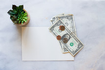 Money, blank paper, and house plant on a white marble counter top. 
