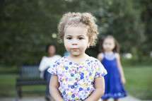 toddler girl standing in front of her family sitting on a park bench 