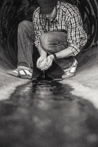 Man cupping flowing water with his hands while kneeling in a sewer drain pipe painted with graffiti.