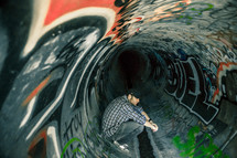 Man sitting ion a sewer drain pipe painted with graffiti.
