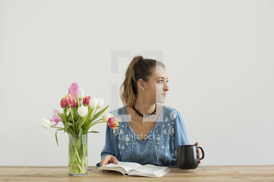 a woman sitting at a desk with a mug of coffee and a vase of tulips reading a book 