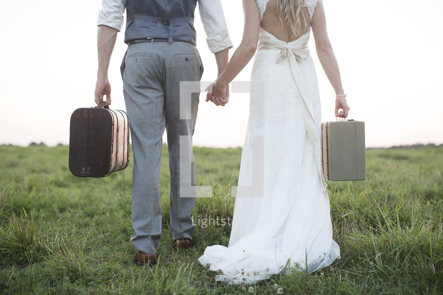 bride and groom carrying luggage in an open field. 