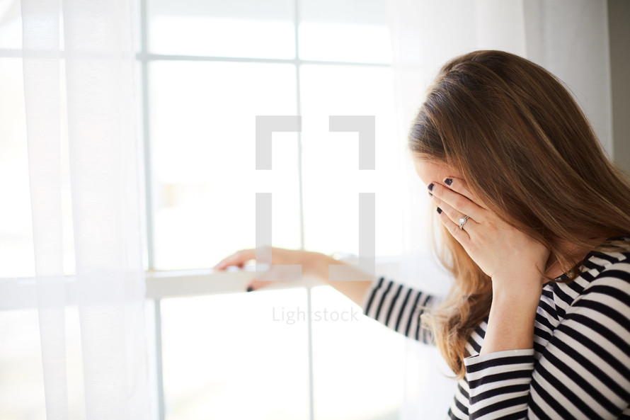 a woman standing in front of a window covering her face with her hand 