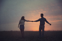 A man and a woman hold hands in a field at dusk.