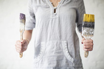 A woman holds a paint brush in each hand.