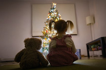 A Little Girl Looking At Christmas Tree With Her Teddy Bear At Home
