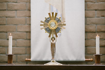 Table with a monstrance and candles in front o a brick wall.