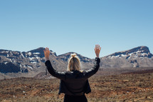 A woman raises her hands to the sky before a mountain range.