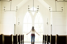 Woman with her arms extended in front of a window while praying in a church.