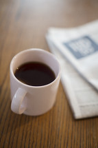 A cup of coffee next to a newspaper on a table.