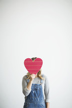 a woman in overhauls holding a red wooden heart 