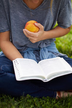 a person sitting in the grass reading a Bible holding a peach 