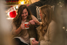Two women sit together drinking hot cocoa from Christmas cups.