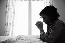 An African American woman kneeling in prayer beside her bed 