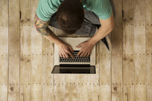 an above view of a man sitting on a wood floor typing on a laptop computer.