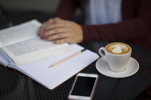 a young woman reading a Bible and journaling at a table with a mug of coffee and cellphone 