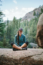 A smiling woman sitting cross legged on a boulder surrounded by a forest.