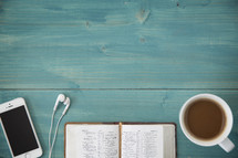 An open Bible, cellphone, ear buds, and cup of coffee on a green wooden table.
