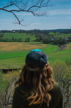a woman standing outdoors looking out at a rural landscape 