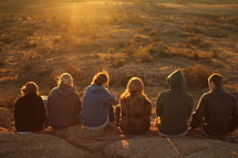 friends sitting at the edge of a cliff in a desert landscape at sunset 