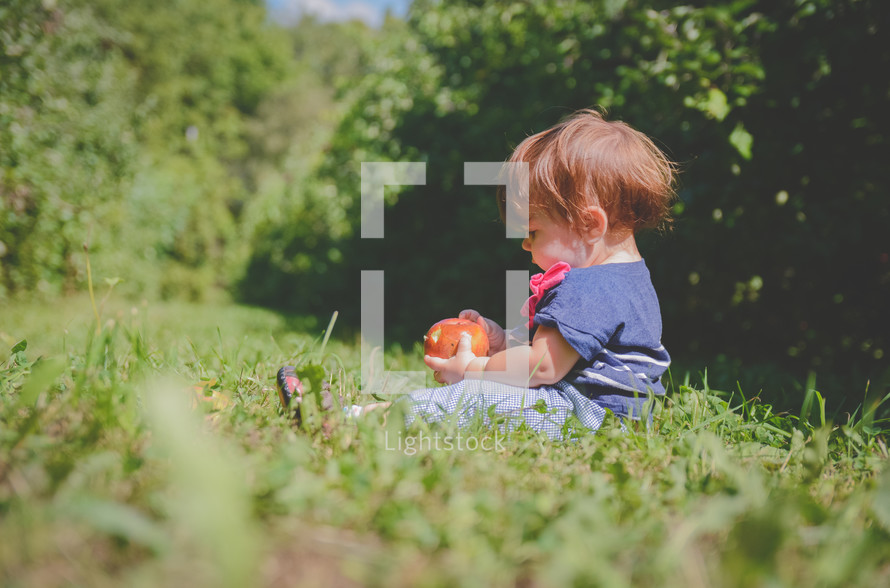 a toddler sitting in grass holding an apple 