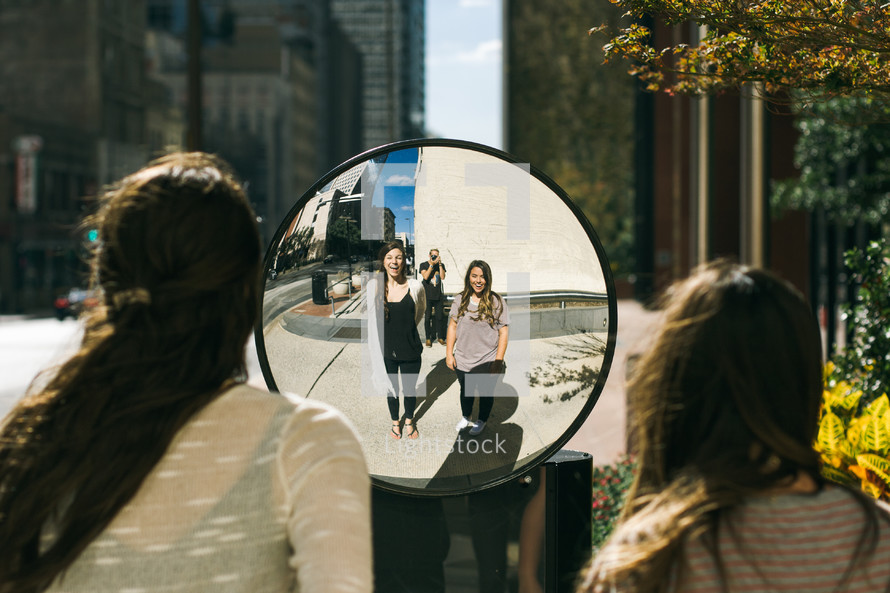 two girl looking into a mirror on a sidewalk 