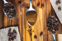 Spoons with coffee powder and coffee beans, laying down on a wooden background.