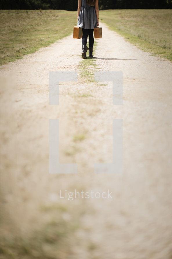 woman walking down a dirt road carrying suitcases 