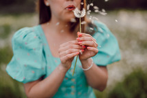 Smiling woman beautiful blowing on ripened dandelion in park. Girl in retro turquoise dress enjoying summer in countryside. Wishing, joy concept. Springtime, aesthetic portrait. High quality photo
