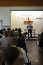 People sitting at tables in a chapel, classroom while a man reads from a Bible at the front