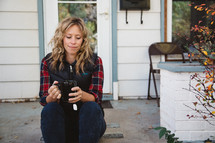 a woman sitting on a stoop in front of her house holding a mug of tea 