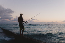 silhouette of a fisherman standing on a rock holding a fishing pole 