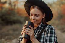 Hipster woman enjoys aroma and taste of fresh coffee. Drinking from thermos in autumn forest. High quality photo