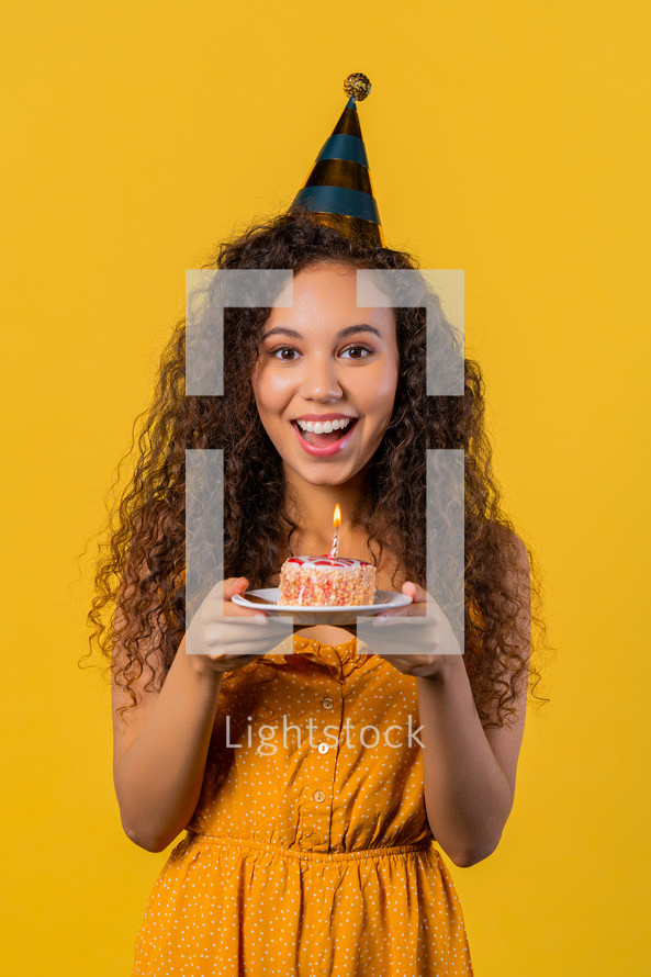 Happy birthday woman making wish - candle on cake. Girl smiling, celebrating anniversary. Young stylish lady on yellow background. High quality photo