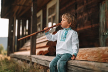 Little boy playing on woodwind wooden flute - ukrainian sopilka. Folk music concept. Musical instrument. Kid in traditional embroidered shirt - Vyshyvanka. Child sitting on porch of old wooden house.