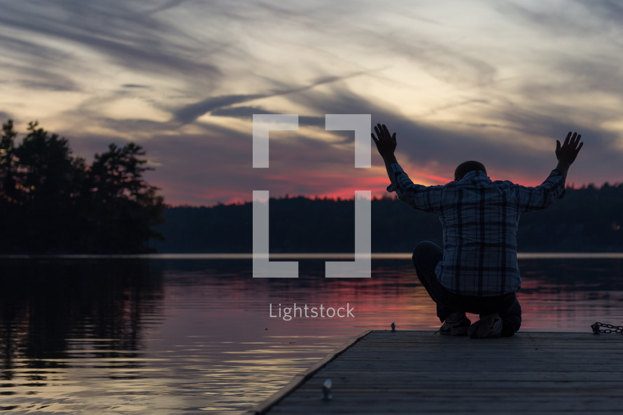 Silhouette of a man with arms raised in praise, on a wooden pier at sunset over a lake.
