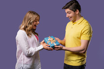 Young american couple on violet studio background. White man gives gift with bow to wife or girlfriend. Birthday, celebration, surprise, anniversary concept. High quality