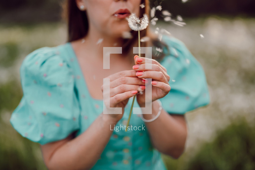 Smiling woman beautiful blowing on ripened dandelion in park. Girl in retro turquoise dress enjoying summer in countryside. Wishing, joy concept. Springtime, aesthetic portrait. High quality photo
